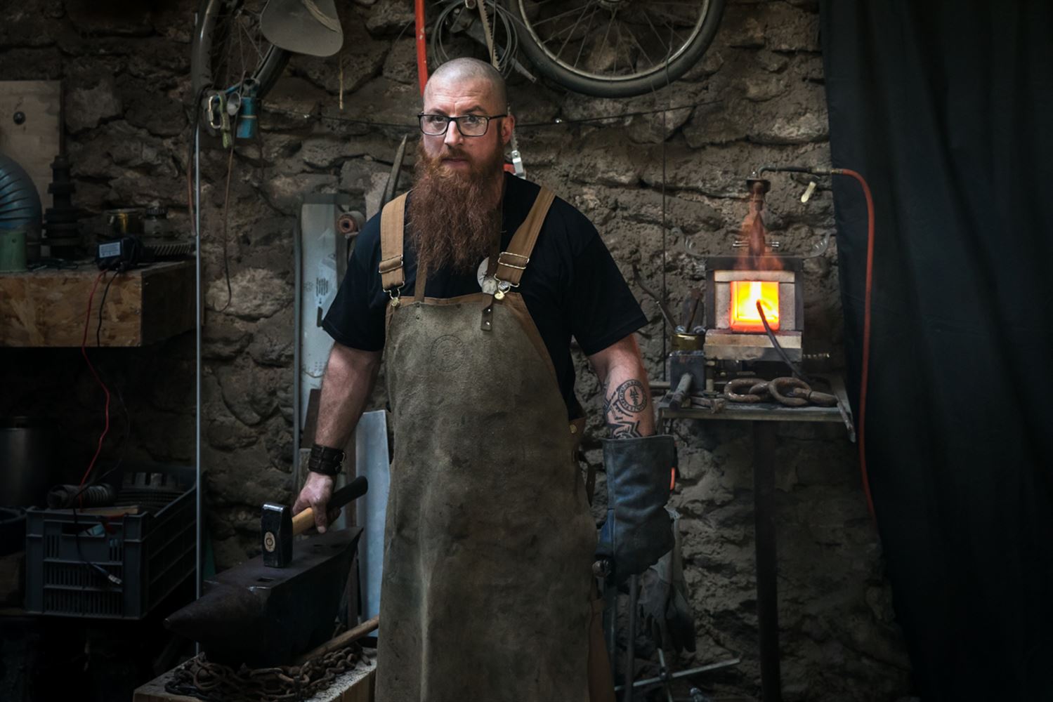 Dans sa forge, Julien à la longue barbe rousse et au crâne rasé pose bravement, la main gauche gantée de cuir, la main droite tient un lourd marteau reposant sur une enclume.